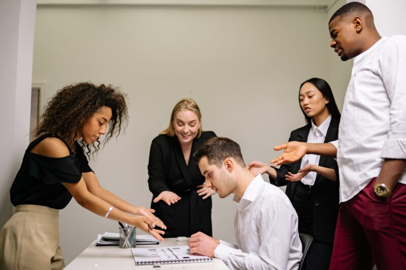 Multiple People Standing and Pointing at a Person Who's Sitting in Front of Documents