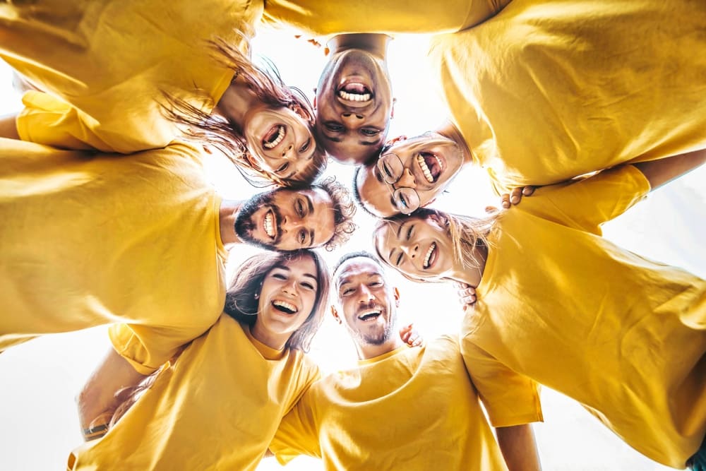 Group Of People With Yellow Shirts On Forming A Circle And Looking Down At The Camera